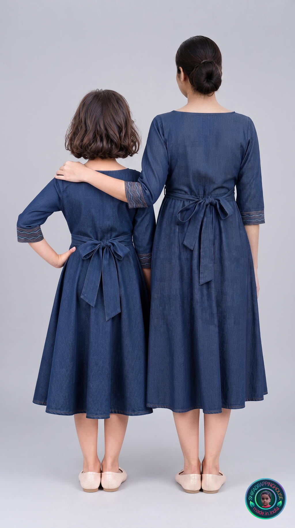 Two women, an adult and a child, standing side by side from the back wearing matching dark blue midi dresses with lace sleeve cuffs and a tie waist. The adult has her arm around the child.