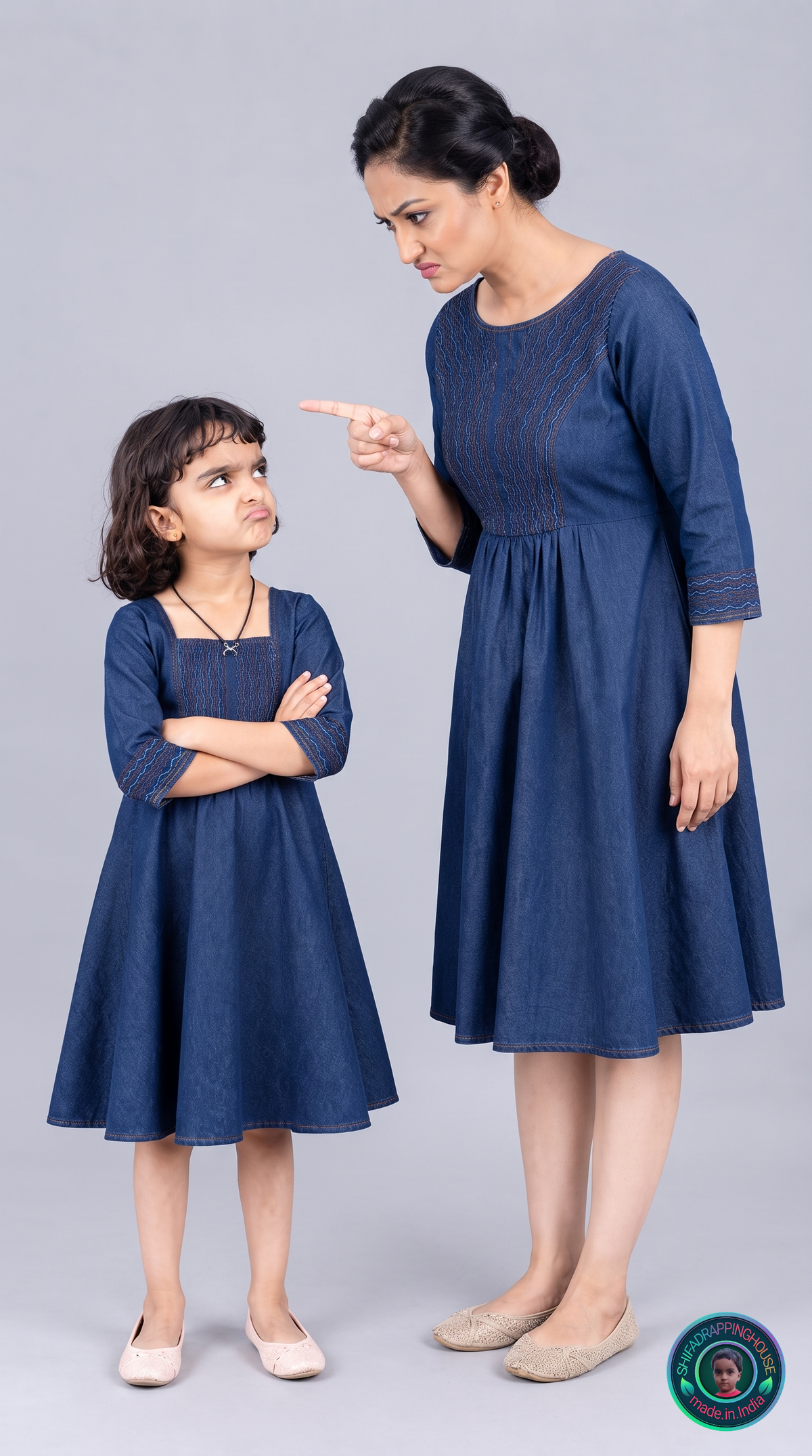 A woman and a young girl wearing matching navy blue embroidered kurta dresses pose against a solid gray studio background. The woman points upward while the girl stands with arms crossed.