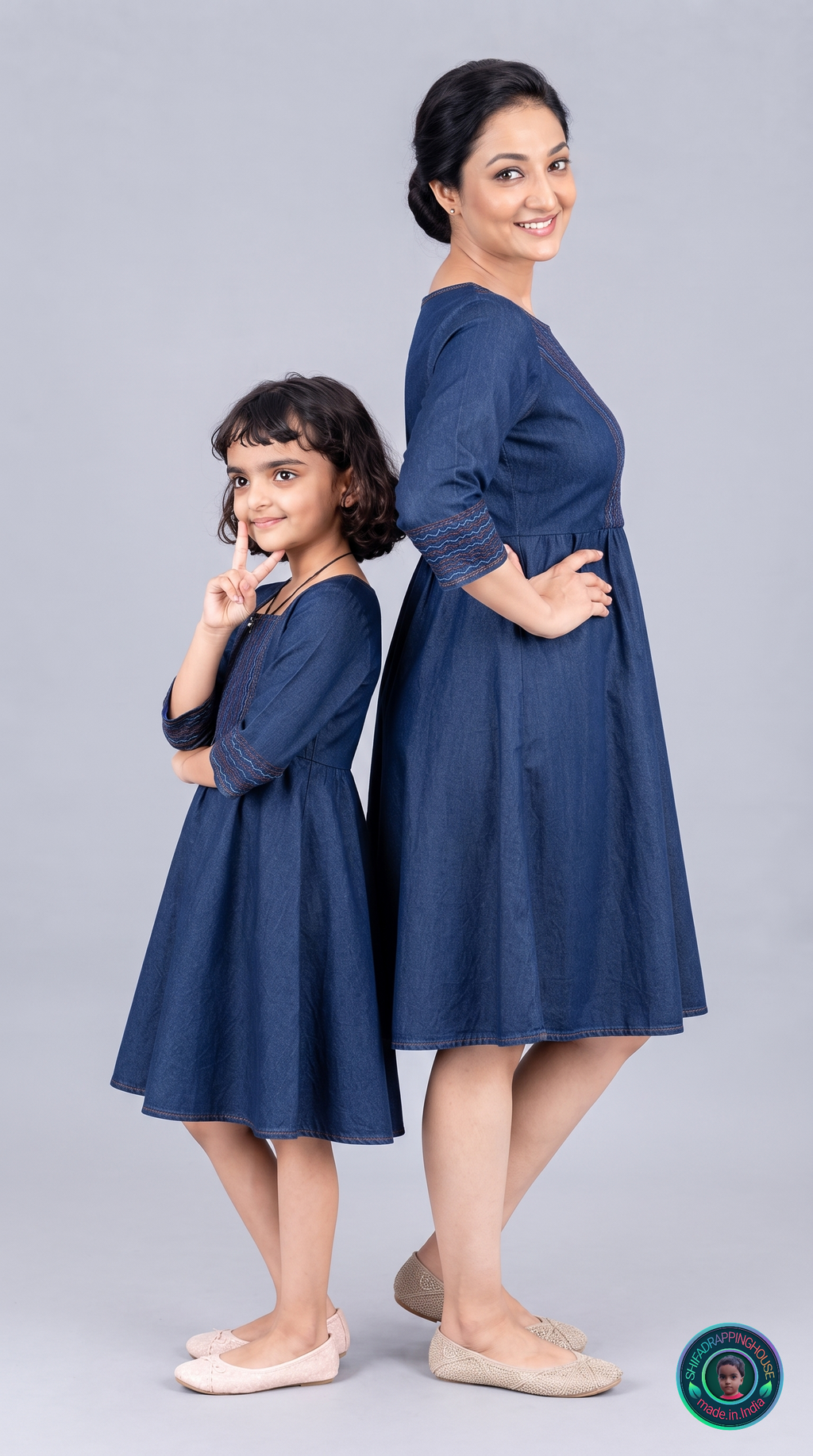 A woman and a young girl modeling matching navy blue dresses with three-quarter sleeves and subtle embroidery on the cuffs, posed back-to-back against a light gray studio background.