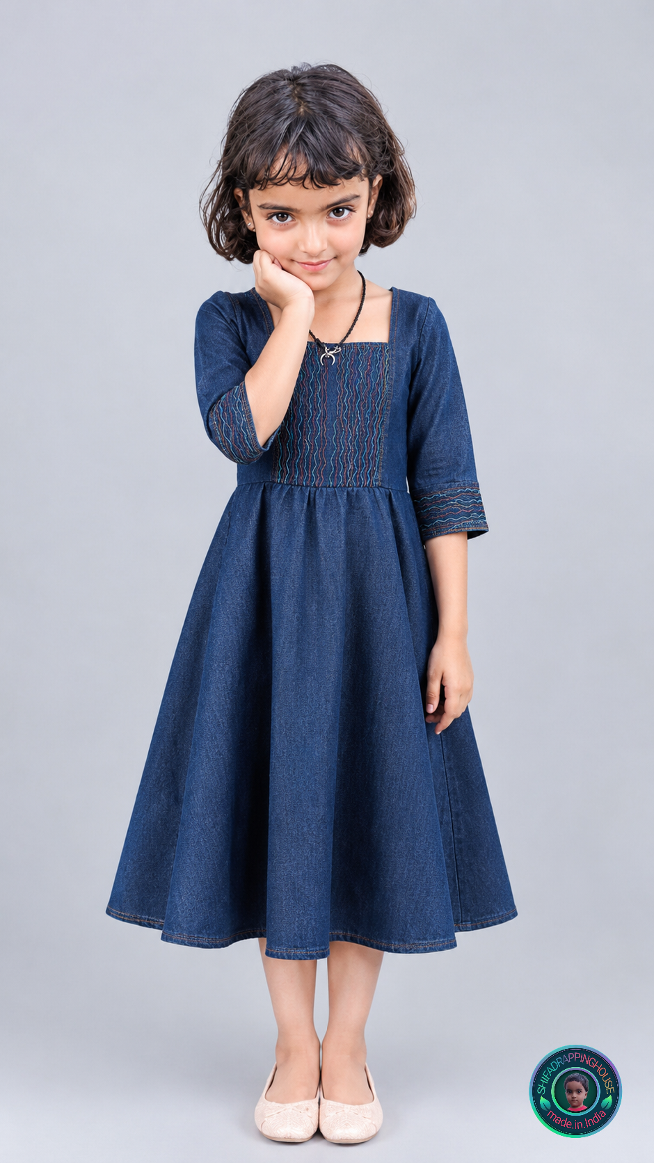 A young girl with dark hair wearing a dark blue denim-style dress with gold embroidery, standing against a solid gray studio background. She is wearing light pink shoes.