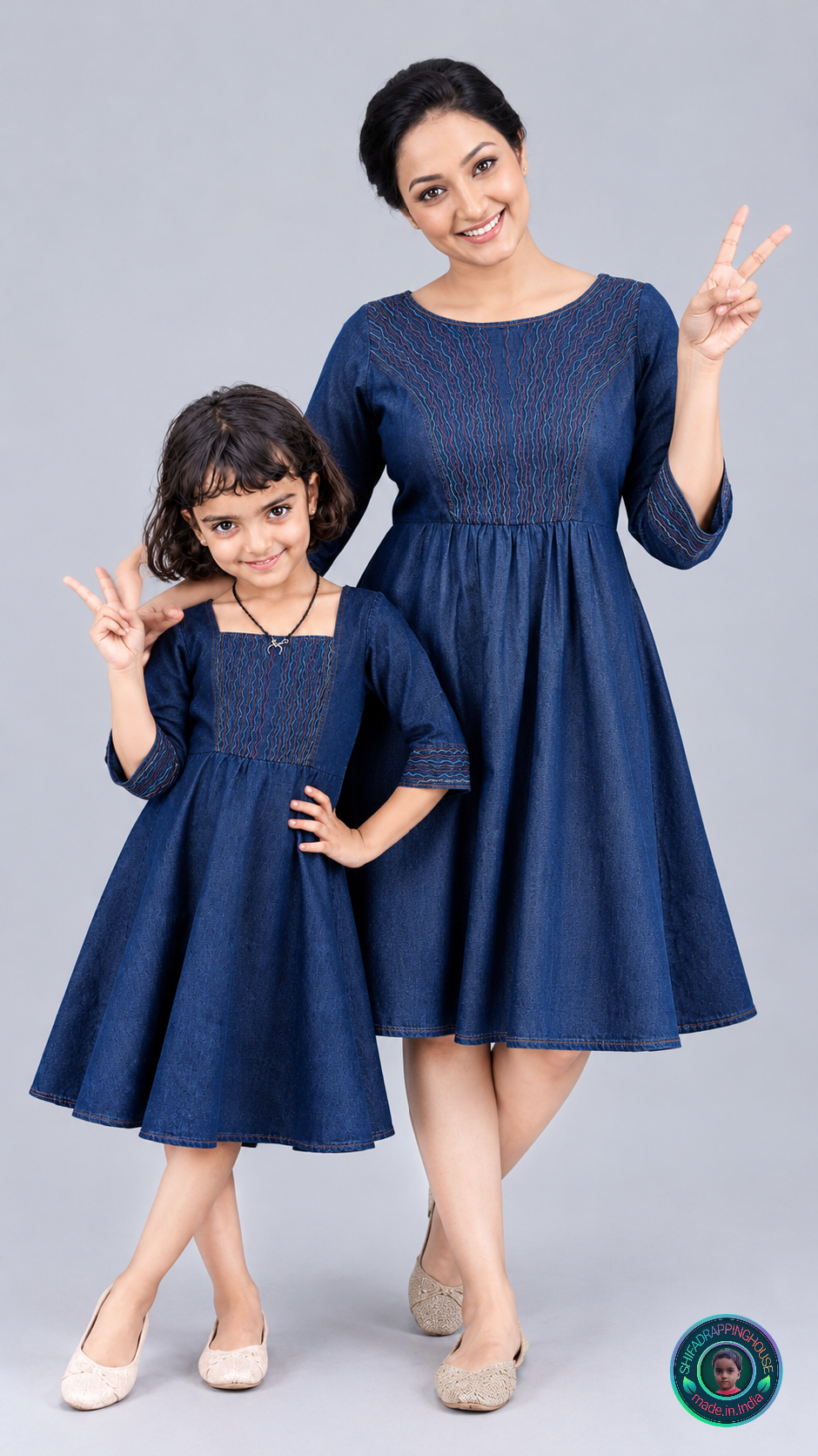 A woman and a young girl modeling matching dark blue denim-style dresses with embroidered bodices, standing against a light gray studio background. Both women are smiling and making peace signs.