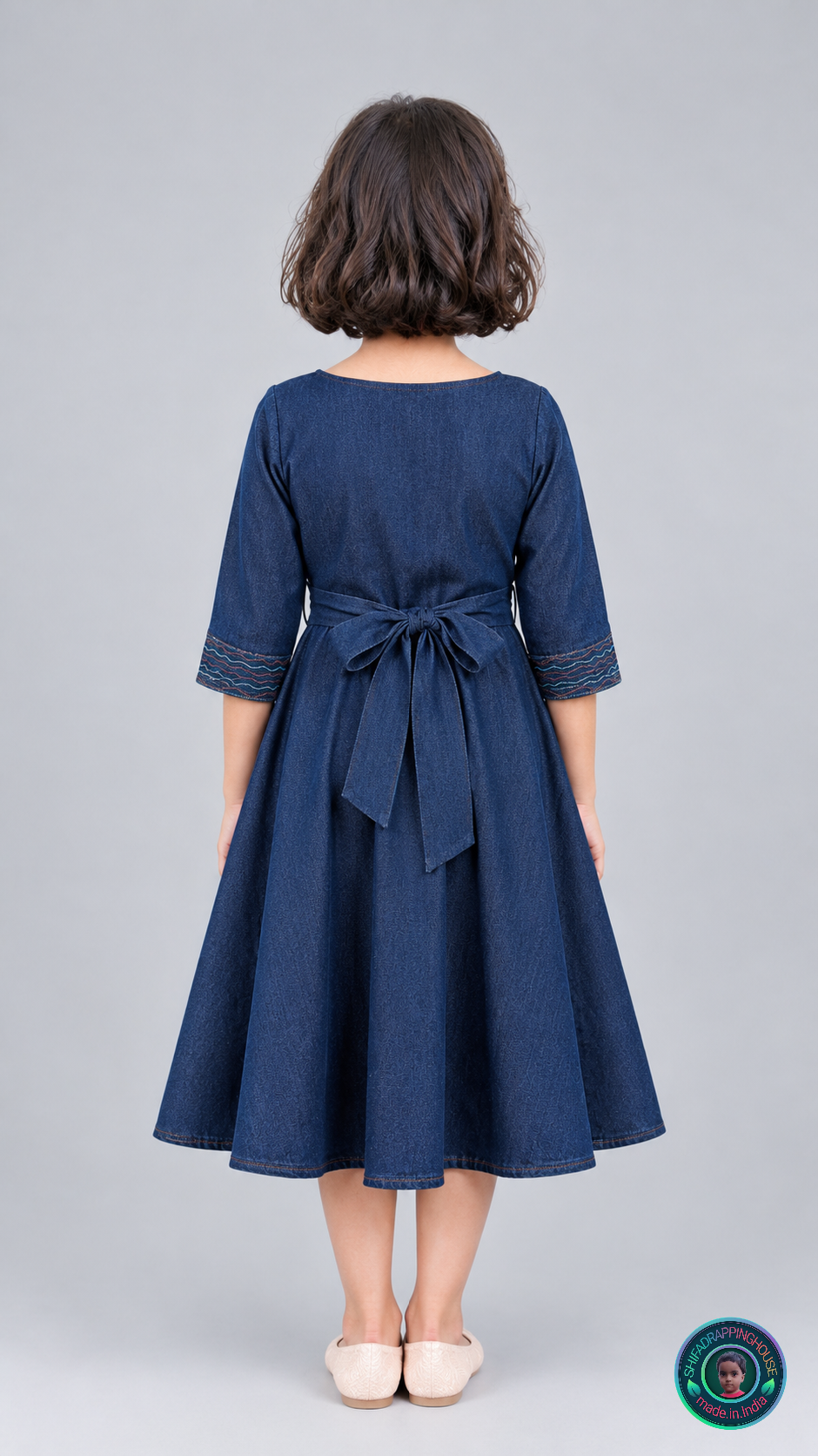 A young girl viewed from behind wearing a dark blue denim-style dress with three-quarter sleeves and a tied waist bow, standing against a light gray studio background.
