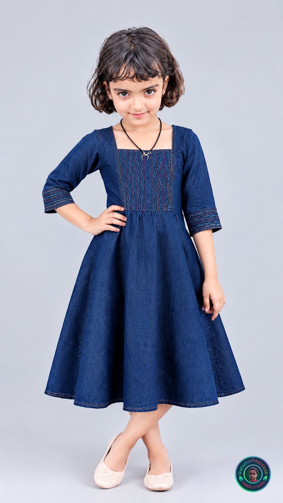 A young girl with short dark hair models a dark blue denim-style dress with three-quarter sleeves and a square neckline, standing against a light gray studio background.