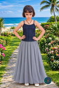 A young girl modeling a two-piece dress with a navy blue and gold striped tank top and a flowing gray skirt, standing on a tropical beach path with the ocean and palm trees in the background.